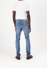 Mäns blå denimjeans med slim fit, standard fem-fickorsdesign, ljus blekning och en läderlapp vid midjan. Bärs med en vit t-shirt.