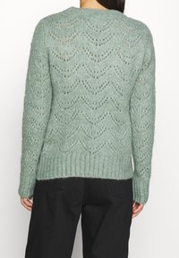 Mintgrön stickad tröja med ett zigzagmönster, med rund halsringning och ribbad nederkant. Materialet verkar mjukt och texturerat.