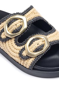 Sandal in materiale naturale tessuto in beige con accenti neri, caratterizzato da due grandi fibbie in tono oro e una suola piatta nera.