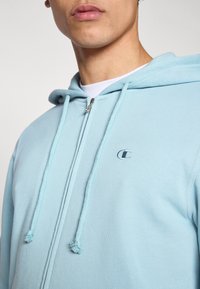 Φωτεινό γαλάζιο φούτερ με φερμουάρ μπροστά και ρυθμιζόμενα κορδόνια. Διαθέτει κεντημένο λογότυπο στην αριστερή πλευρά του στήθους. Μαλακή υφή υφάσματος.