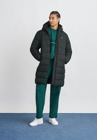 Νεαρός άνδρας στέκεται με τα χέρια στις τσέπες, φορώντας μαύρο μακρύ μπουφάν, πράσινη μπλούζα και παντελόνι, και λευκά αθλητικά παπούτσια πάνω σε μπλε χαλί.