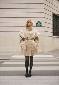 Szőke nő bézs színű ballonkabátot, fekete harisnyát és cipőt viselve áll egy városi gyalogátkelőhelyen, egy kőépület előtt.