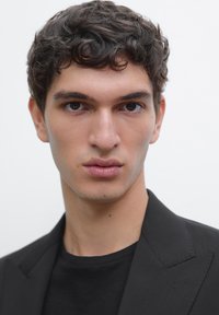 Porträt einer Person mit dunklem, lockigem Haar, die einen schwarzen Blazer über einem schwarzen T-Shirt trägt. Der helle Hintergrund verstärkt den Kontrast zur Kleidung.