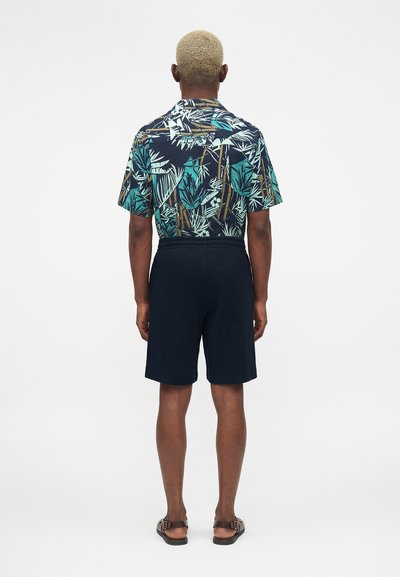 Άνδρας με κοντά ξανθά μαλλιά, που φοράει μαύρο, μπλε και πράσινο πουκάμισο με τροπικό μοτίβο, μαύρα σορτς και καφέ σανδάλια, στέκεται με την πλάτη στραμμένη.