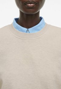 Beżowa bluza z okrągłym dekoltem, fakturowanym materiałem i wytłoczonym logo. Jasnoniebieska koszula z kołnierzykiem pod spodem.