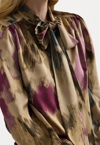 Žena v hedvábné blůze s nafouklými rukávy, uvázanou mašlí u límce a abstraktním vzorem v béžové, fialové, modré a hnědé barvě.