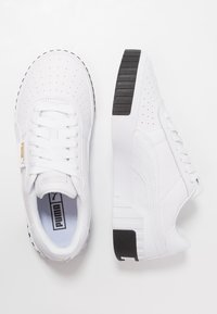 Zapatillas blancas con textura perforada, cordones blancos planos y una suela negra con patrón. El lado presenta un logo sutil y un acabado de cuero suave.