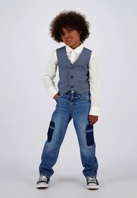 Svijetloplavi prsluk preko bijele košulje, uparen s plavim trapericama s jastučićima na koljenima. Crno-bijele tenisice zaokružuju odjevnu kombinaciju.