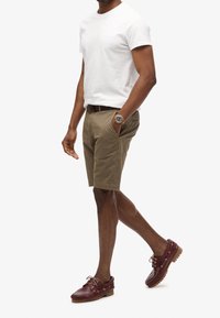 Pánský outfit skládající se z bílého trička, khaki šortek a vínových lodních bot s hnědými tkaničkami. Doplňky zahrnují zlaté hodinky.