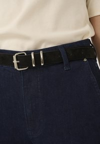 Sort ruskindsbælte med sølvmetalspænde, bæret over en lysfarvet skjorte og mørkeblå denimbukser, med en glat tekstur.