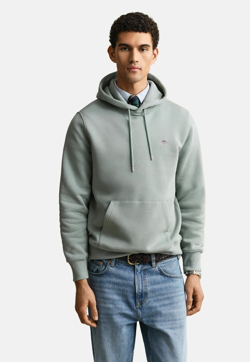 Giovane uomo che indossa una felpa con cappuccio grigio chiaro sopra una camicia con colletto e cravatta a righe, abbinata a jeans blu e un orologio da polso.