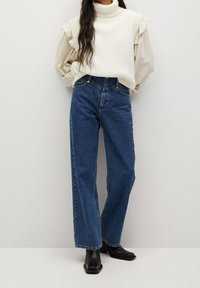 Jeans en denim bleu taille haute avec une coupe droite, associés à un col roulé côtelé crème et des manches superposées. Bottines noires à bouts carrés.