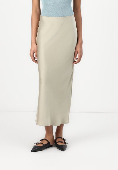 Jupe en satin vert olive clair avec une texture lisse, taille ajustée et une silhouette fluide. Associée à des ballerines noires à bouts pointus ornées de deux nœuds.