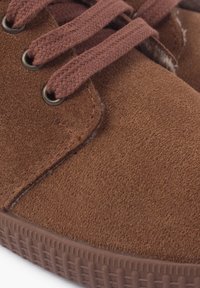 Chaussure en suede marron avec bouts ronds et surface texturée. Dispose de lacets marron enfilés à travers des œillets en métal. Semelle en caoutchouc avec motif antidérapant.