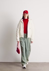 Weißer Reißverschluss-Fleecejacke, roter Rollkragenpullover, hellgrüne weite Jeans, rote Handtasche und rote Mütze, kombiniert mit weißen Turnschuhen. Einfarbiger Hintergrund.