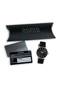 Montre OOZOO noire avec aiguilles et index argentés, bracelet en cuir et boucle réglable, présentée aux côtés d'une carte de garantie noire et de son emballage.