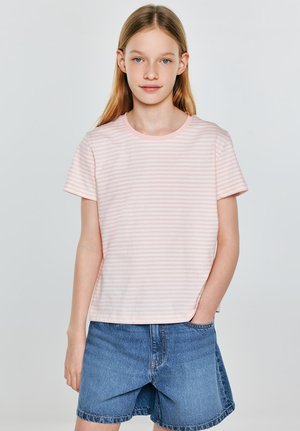 Noor inimene, kellel on pikad blondid juuksed, kannab roosaid ja valgeid triibulist t-särki ning siniseid teksaseid lühikesi pükse, seisab ühe käega taskus.