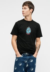 Νεαρός άνδρας με κοντά καστανά μαλλιά, φορώντας μαύρη μπλούζα Santa Cruz και μπλε τζιν με σύμβολα yin-yang, με τα χέρια στις τσέπες, κοιτάζοντας στο πλάι.