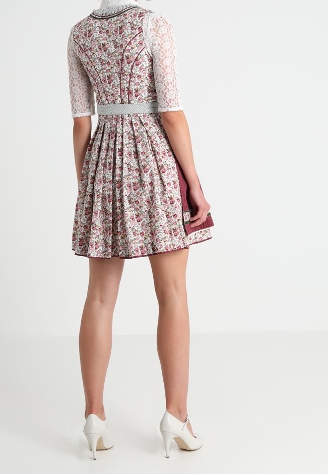 Femme portant une robe à motif floral avec des manches en dentelle et des talons hauts blancs, debout devant un fond clair uni.