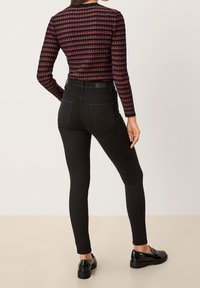 Žena v černých skinny džínech, s dlouhým pruhovaným topem v černé a červené barvě a černými mokasínami, stojící zády k fotoaparátu.