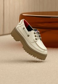 Κρεμ δερμάτινο παπούτσι σκαφού με κόκκινη εσωτερική επένδυση και χοντρή καφέ σόλα με αυλακώσεις, τοποθετημένο σε μπεζ χαλί με θολό καφέ τραπέζι στο φόντο.