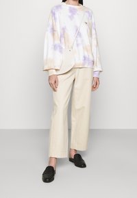 Person som bär krämfärgade vida byxor, svarta slip-on skor, en pastellfärgad tie-dye sweatshirt och en liten axelremsväska mot en enfärgad bakgrund.