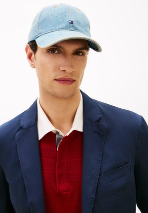 Fiatal férfi világoskék farmer sapkával, tengerészkék blézerrel, fehér galléros inggel és piros kötött polóval egy stúdióban.