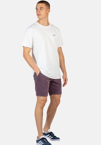 Camiseta blanca de manga corta, pantalones cortos morados y zapatillas azules. La camiseta presenta un pequeño logo, los pantalones cortos tienen una tela texturizada. Atuendo casual.