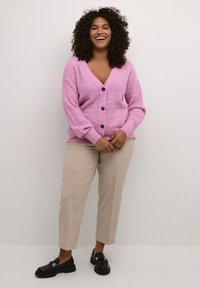 Cardigan roz, texturat, cu decolteu în V, mâneci lungi și cinci nasturi negri în centru; asortat cu pantaloni bej și mocasini negri.