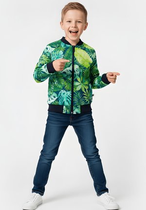 Zielona kurtka bomber z tropikalnym wzorem liści, granatowymi akcentami, zamkiem błyskawicznym, w połączeniu z ciemnymi dżinsami i białymi trampkami.