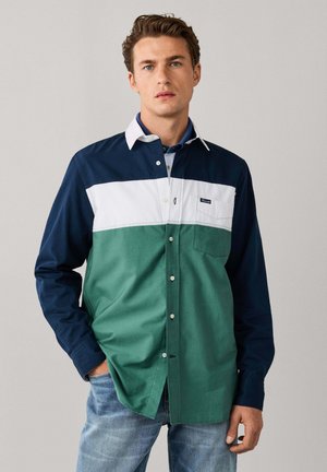 Hombre con camisa de manga larga con bloques de colores azul marino, blanco y verde y vaqueros azul claro, de pie con una mano en el bolsillo.