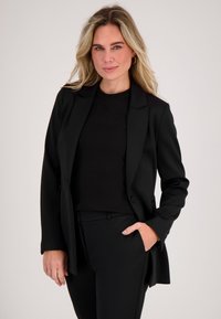 Blonde vrouw die een zwarte blazer, zwarte top en zwarte broek draagt, staand met één hand in de zak tegen een eenvoudige lichte achtergrond.