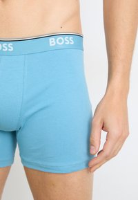 Hellblaue Boxershorts aus weichem Stoff, mit einem weißen Logo-Bund und schwarzen sowie goldenen Akzenten. Taillierter Schnitt und glatte Textur.