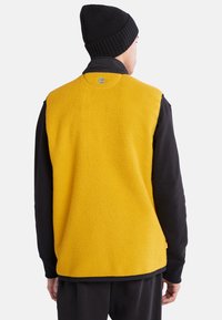Fleece vest i lys gul med teksturert overflate, med en svart langermet skjorte under og en svart strikket lue.