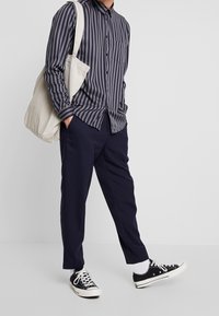 Camicia a righe blu navy con bottoni, taschino sul petto, abbinata a pantaloni blu navy. Borsa a tracolla in tela e sneakers nere con suola bianca.