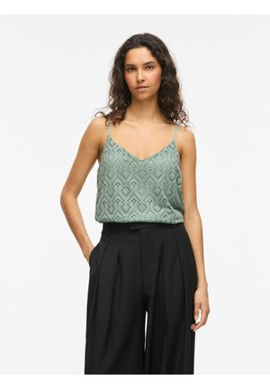 Jeune femme aux cheveux bouclés portant un haut sans manches à motif vert clair et un pantalon noir plissé taille haute.