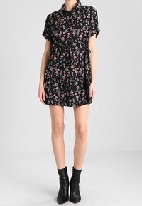 Schwarzes Blumenkleid mit kurzen Ärmeln, Knopfleiste, tailliertem Schnitt und verstreuten roten und weißen Blumenmustern. Getragen mit schwarzen Knöchelstiefeln.