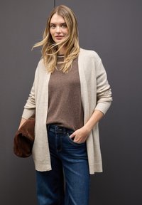 Cardigan beige sopra un maglione marrone e jeans blu scuro, con una vestibilità comoda. Il cardigan ha polsini a coste e un design semplice.