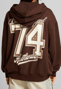 Hnědá mikina s kapucí, na zadní straně s velkým bílým a béžovým grafickým designem "74" a "Authentic", s uvolněným střihem a šňůrkou na kapuci.