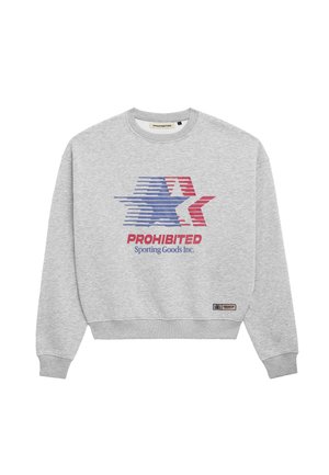 Młoda kobieta w granatowej czapce i szarej bluzie z czerwono-niebieską gwiazdą w paski oraz napisem „PROHIBITED Sporting Goods Inc.” stoi na białym tle.
