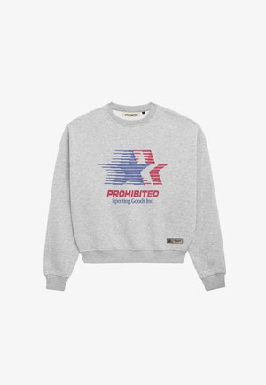 Młoda kobieta w granatowej czapce i szarej bluzie z czerwono-niebieską gwiazdą w paski oraz napisem „PROHIBITED Sporting Goods Inc.” stoi na białym tle.