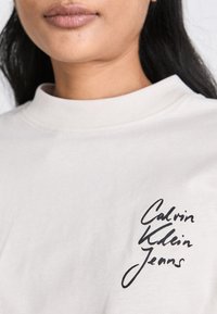 T-shirt beige à col rond avec un logo noir manuscrit "Calvin Klein Jeans" sur le côté gauche de la poitrine. Tissu en coton lisse, coupe décontractée.