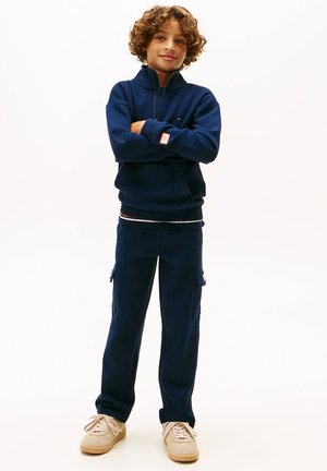 Sweatshirt zippé bleu marine avec un motif texturé, associé à un pantalon cargo de style assorti. Des chaussures décontractées de couleur claire complètent la tenue.