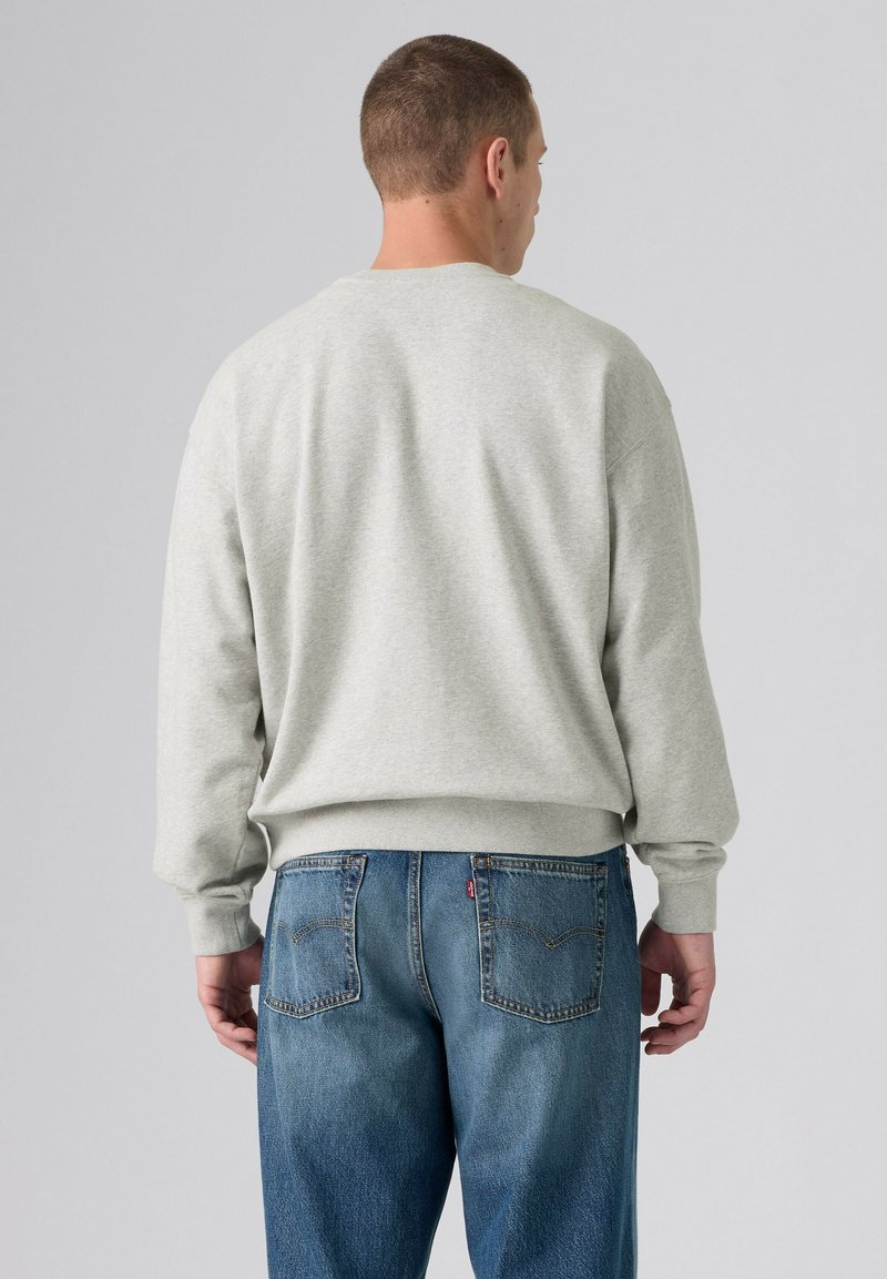 Uomo che indossa una felpa grigio chiaro e jeans blu, visto di spalle su uno sfondo grigio chiaro uniforme.