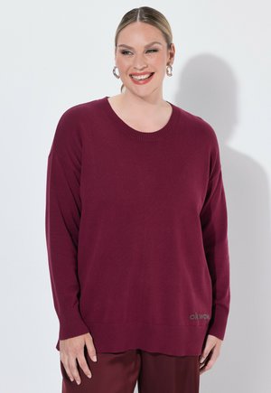 Smilende kvinde med lyst hår bundet tilbage iført store øreringe og en løs mørkerød sweater og matchende bukser mod en ensfarvet baggrund.