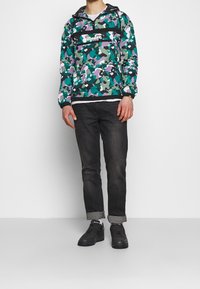 Camouflage pulloverjas in groen, paars en zwart; voorzak met logo; gecombineerd met donkere jeans en zwarte schoenen.
