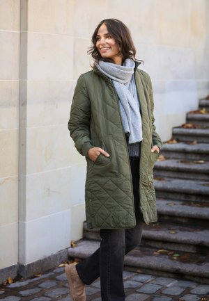 Olivgrün gesteppter langer Mantel mit Reißverschluss vorne, Seitentaschen und einem weichen grauen Schal. Schwarze Jeans und beigen Stiefel runden das Outfit ab.