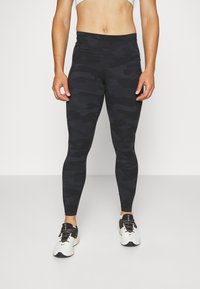 Juodos kamufliažo leggings'ai su aukštu liemeniu ir lygiu paviršiumi, derinami su baltais ir juodais sportiniais batais, turinčiais tekstūruotą dizainą.