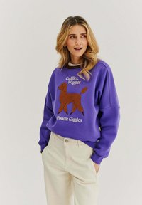 Fialová mikina s grafikou hnědého pudla a texty "Cuddles, Wiggles" a "Poodle Giggles" v bílé barvě. Měkká tkanina, uvolněný střih.