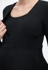 Κοντινό πλάνο ενός ατόμου που φοράει μαύρη μπλούζα τύπου wrap με στρογγυλή λαιμόκοψη και μακριά μανίκια, ρυθμίζοντας το ύφασμα κοντά στο στήθος.